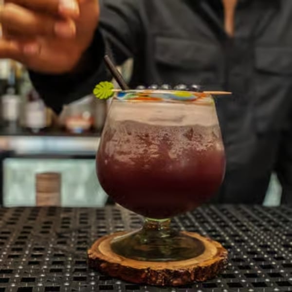 A bartender's hand reaching for a purple cocktail with foam, garnished with berries and a tropical pick.