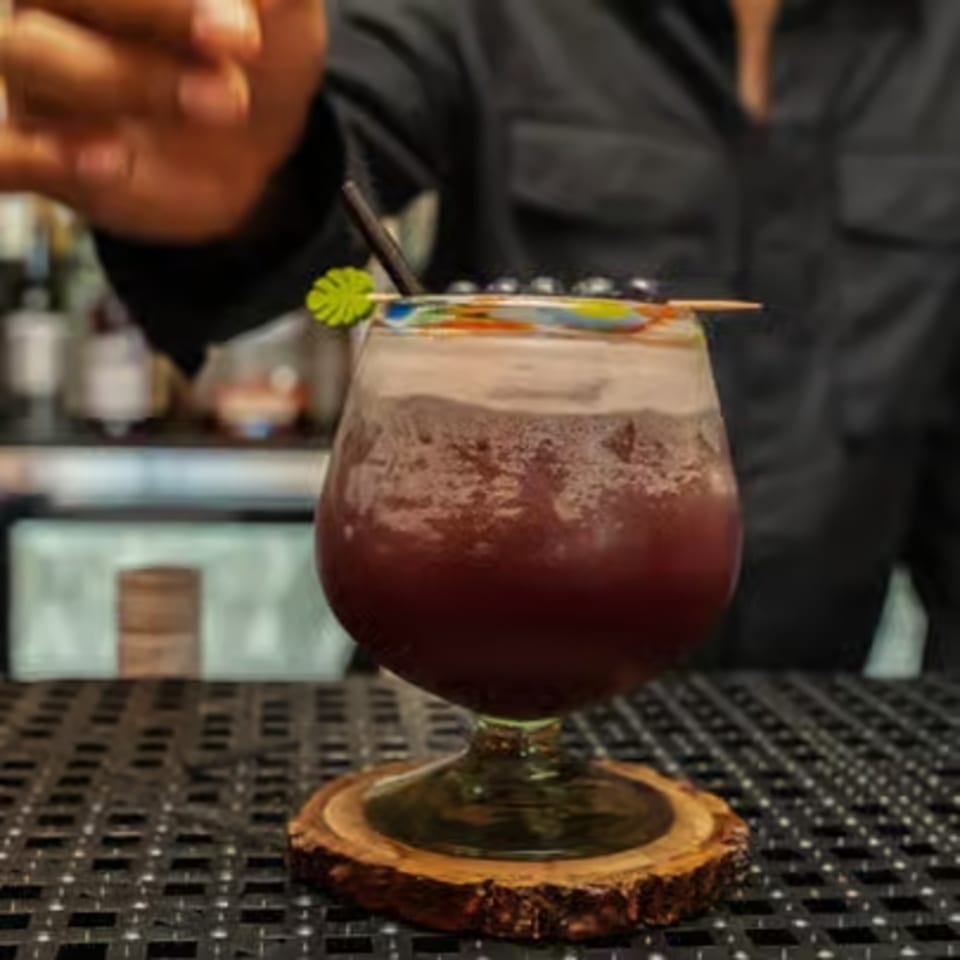 A bartender's hand reaching for a purple cocktail with foam, garnished with berries and a tropical pick.