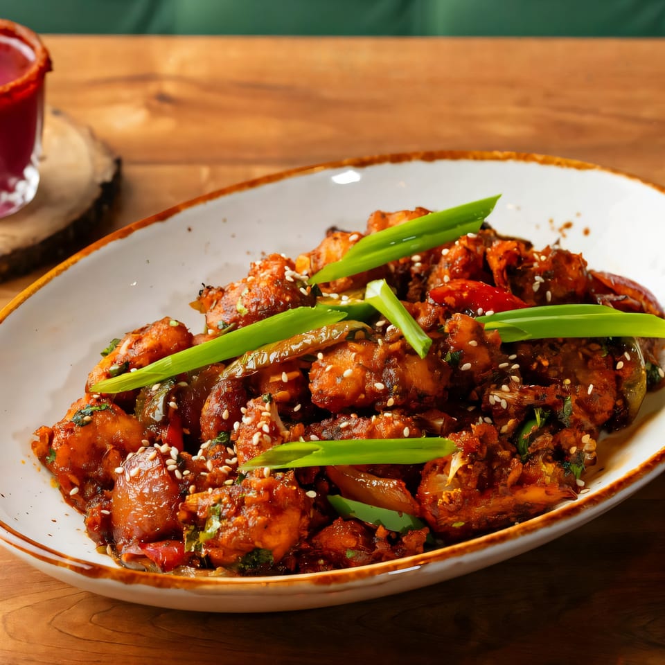 A plate of Indian chili chicken, garnished with sesame seeds and green onions, on a wooden table.