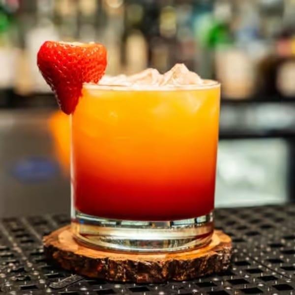 A vibrant orange and red cocktail, garnished with a fresh strawberry, sits on a wooden coaster.