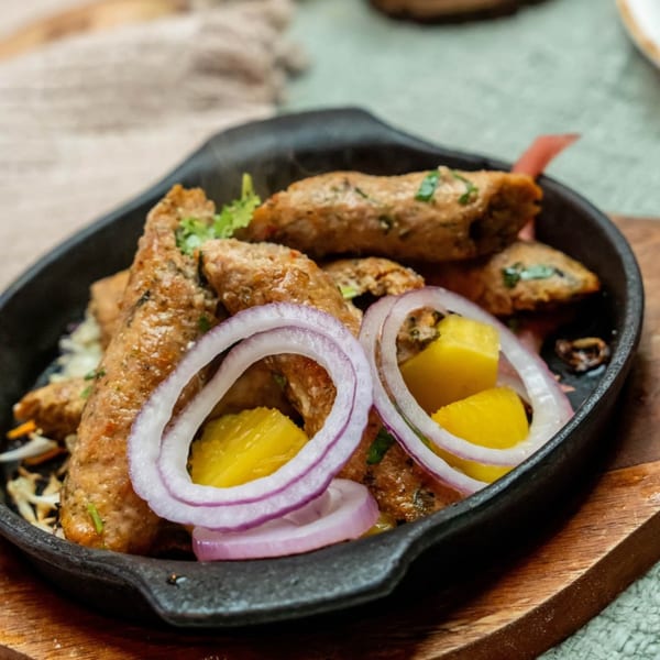 Indian seekh kebabs with red onion rings and pineapple chunks in a black cast iron dish.