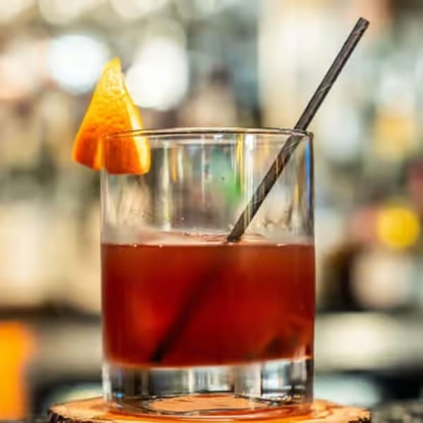 A red cocktail with an orange slice and black straw, on a wooden coaster.